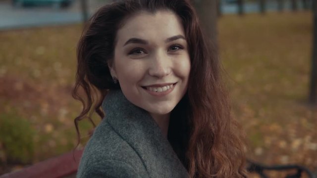 Young brunette girl in romantic look, with curly hairstyle in coat sitting at bench, looking at camera and smiling in slow motion. Park and trees on background.