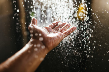 A man's hand in a spray of water in the sunlight against a dark background. Water as a symbol of purity and life - obrazy, fototapety, plakaty