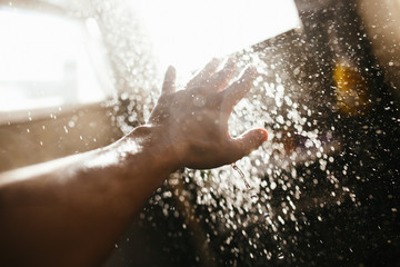 A man's hand in a spray of water in the sunlight against a dark background. Water as a symbol of purity and life