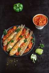 Fried fish in a plate with  tomato sauce, greens and peppers on dark background