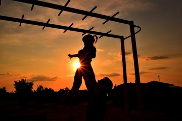 father raises high his little daughter in the hands of the children's playground is the bright evening sun