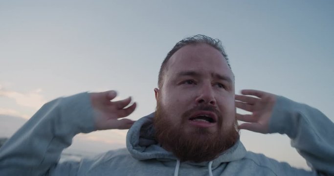 young overweight runner man resting takes off hood exhausted after intense running exercise training difficult weight loss challenge on seaside at sunrise close up