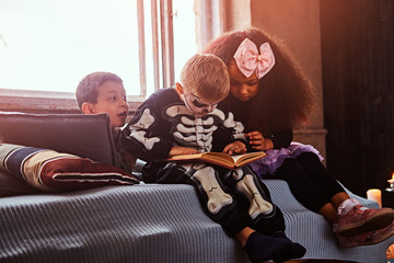 Three multiracial kids in scary costumes reading horror stories while sitting on bed in an old house.