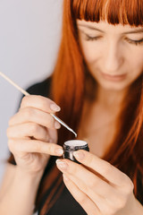 red-haired woman holds a brush with a jar and looks at the texture of gel-lacquer. Close-up