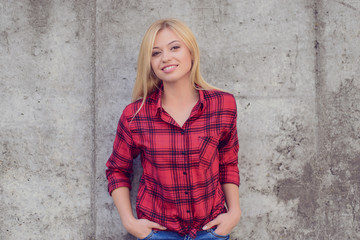 Woman smiling at camera. Woman with blonde hair, dressed in red checkered shirt and jeans standing against grey wall. Portrait, close up photo