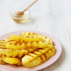 Grilled pineapple wedges on a pink plate with honey over white wooden surface, close-up. Selective focus.