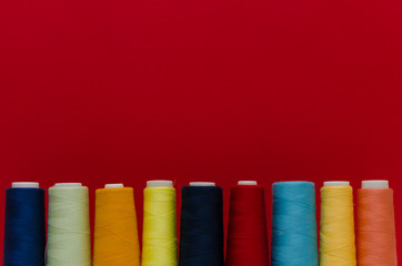 The thread spools of different colors stand in a row on the white wooden table in front of the red background 
