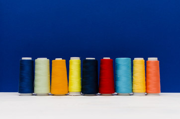 The thread spools of different colors stand on the white wooden table in front of the blue background 
