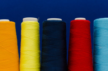 Close-up of the thread spools of different colors stand in a row on the white wooden table in front of the blue background 