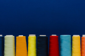 The thread spools of different colors stand in a row on the white wooden table in front of the blue background 