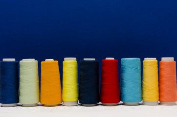 The thread spools of different colors stand on a wooden table in front of the blue background 