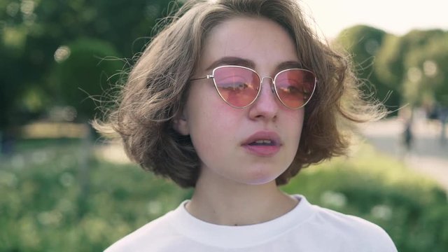Young woman with wavy fair hair wearing pink sunglasses and t shirt smoking cigarette and drinking coffee in summer park. Concept of addiction and bohemian lifestyle. Slider slow motion portrait shot