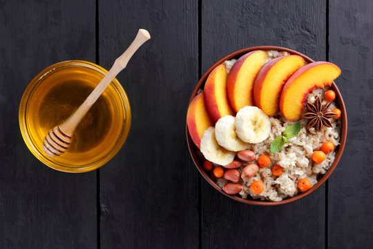 Oatmeal and honey on dark wooden boards. Cooked oatmeal with fruit and honey for breakfast. Healthy food in earthenware. Peaches, bananas, mint and mountain ash with porridge