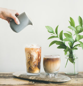 Iced coffee in tall glasses with milk pouring from pitcher by hand, white wall and green plant branches at background, copy space, square crop. Summer refreshing beverage ice coffee drink concept