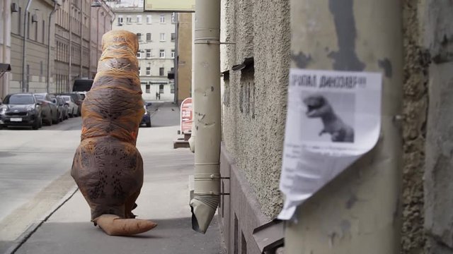 Person in t rex mascot walking away from camera, metal downspout with poster sais DINOSAUR LOST in russian at city street sidewalk