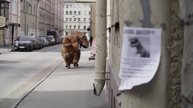 Person in t rex mascot running towards camera, metal downspout with poster sais DINOSAUR LOST in russian at city street sidewalk