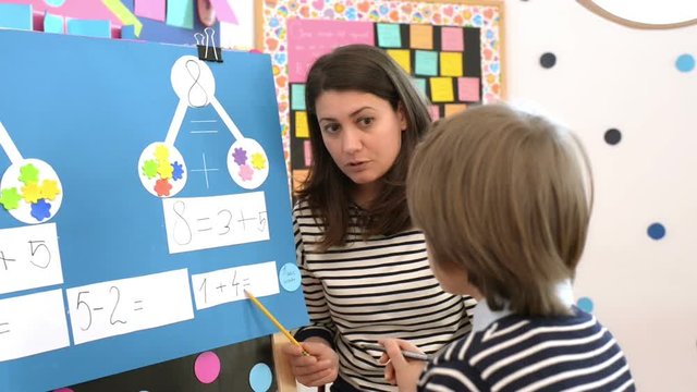 Cute boy solving graphic math exercises at kindergarten, under educator observation