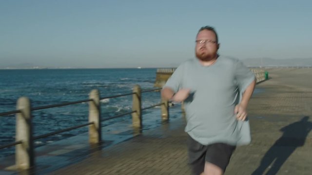 overweight man running intense workout challenge obese caucasian male jogging exercising seaside at sunrise wearing glasses close up