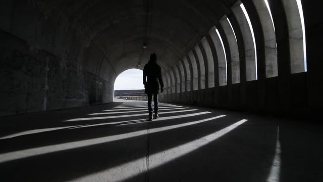 Hand held camera woman walking over street going out a brutalist concrete tunnel. Lights and shadows rithms made by windows, openings, within the tunnel. Flares of sunlight. Argentina, Mendoza