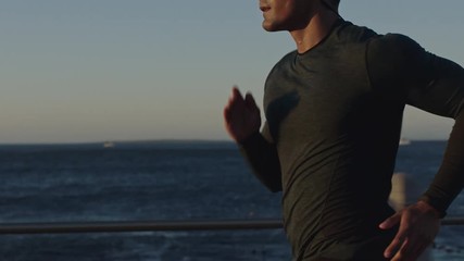 attractive young man running athlete endurance runner training jogging cardio exercise workout on calm ocean seaside run wearing earphones at sunset slow motion - Powered by Adobe