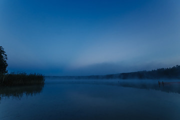 Fog over the lake, twilight over the lake, very dense fog, dawn, blue sky over the lake, the morning comes, the forest reflects in the water, surface water, clear morning sky, gothic, Grim picture