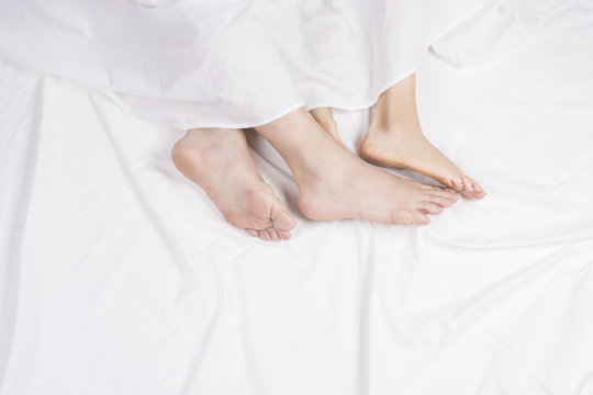 Feet of couple sleeping side by side in comfortable bed. Close up of feet in a bed under white blanket. Bare feet of a man and a woman peeking out from under the cove