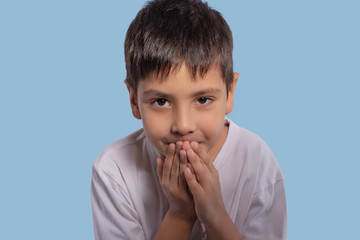 The  little boy wearing a white shirt on blue background in studio. He  is   wants to share a secret, so he leaned forward and covered his mouth with his hands to whisper a secret