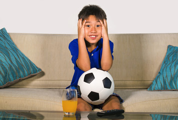 lifestyle portrait at home of young 7 or 8 years old boy holding soccer ball watching excited and nervous football game on television sitting at living sofa couch