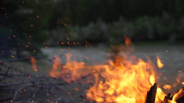Vivid burning bonfire close up at evening. Slow motion, 180fps. Fire burning on the background of the river. Sparks flying from the fire.