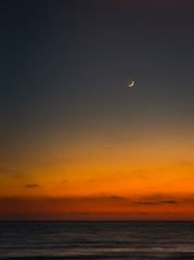 As the sun goes down and seems to melt into the ocean, the skies darken a bit and the moon begins to shine in the seaside sky as friends and lovers sit and enjoy the scene and each other