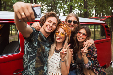 Group of excited hipsters men and women smiling, and taking selfie on mobile phone near vintage minivan into the nature
