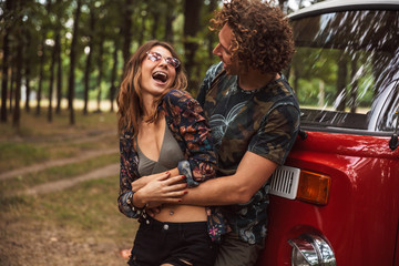 Caucasian hippy couple man and woman smiling, and hugging each other while standing near minivan in...