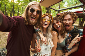 Image of cheerful hippie people men and women, taking selfie in forest near retro minivan