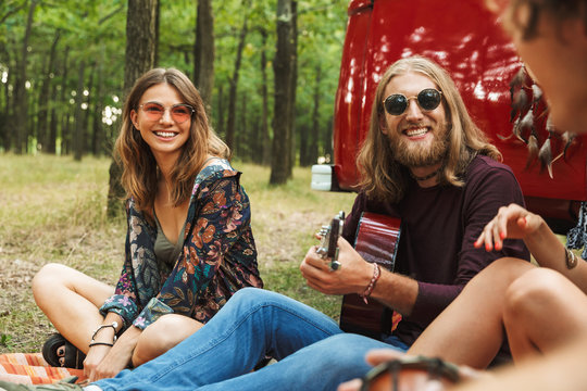 Group of cheerful friends hipsters men and women smiling, and sitting near vintage minivan in forest