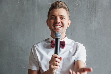 A young attractive funny joyful emotional man winks in a white shirt and a butterfly with a microphone in his hands, on a gray background