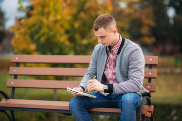 A cute student sits on the bench and writes down his thoughts in his notebool using a pensil. study outside