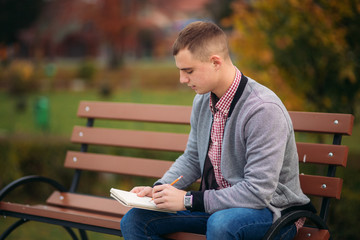 Fototapeta premium A cute student sits on the bench and writes down his thoughts in his notebool using a pensil. study outside