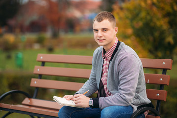 Fototapeta premium A cute student sits on the bench and writes down his thoughts in his notebool using a pensil. study outside