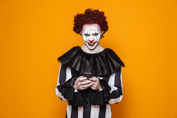 Scary clown man 20s wearing black costume and halloween makeup using smartphone, isolated over yellow background