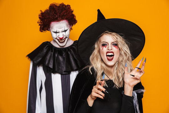 Image of witch woman and joker man wearing black costume and halloween makeup looking at camera, isolated over yellow background