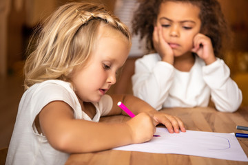 african american kid looking at caucasian child drawing at table in kindergarten