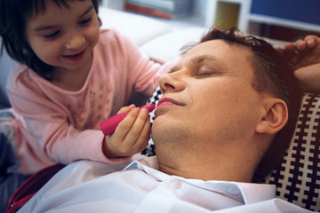 Fototapeta premium Cute little girl apply lipstick to father while he is sleeping. Spending time happily.
