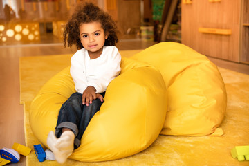 adorable african american kid sitting on bean bag chair in kindergarten and looking at camera