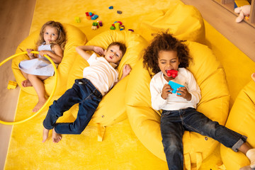 high angle view of multiethnic kids lying on bean bag chairs with toys in kindergarten