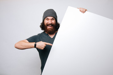  Young bearded man with grey hat hiding behind white sheet and pointing on it on white background.