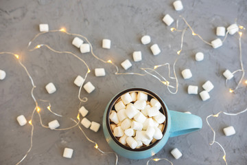 Blue cup of coffee on gray concrete background with marshmallow and garland. Cozy drink autumn and winter concept. Top view. Flat lay.