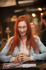 Red-haired young woman in glasses sits in a bar with a mug of beer