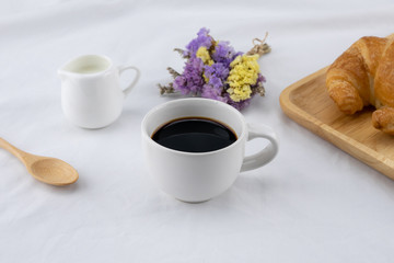 Fototapeta na wymiar A cup of coffee, milk and croissant with statice flower bouquet on white bed, selective focus on coffee