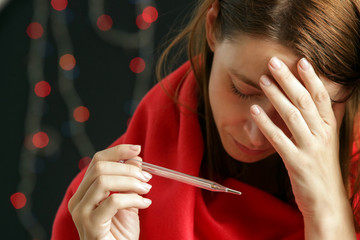 Young woman wrapped in warm red coverlet holds thermometer which shows high temperature, cold and...