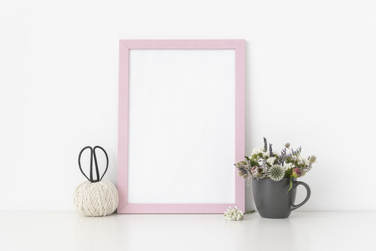 Pink a4 portrait frame mockup with dried field wild flowers in gray mug and vintage scissors on white wall background. Empty frame, poster mock up for presentation design. Template frame for text